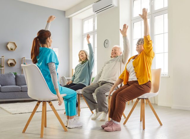 Young nurse leading seated exercise session with elderly patients in nursing home. Group of happy senior people sitting on chairs, raising their arms during physical therapy for mobility and wellness.