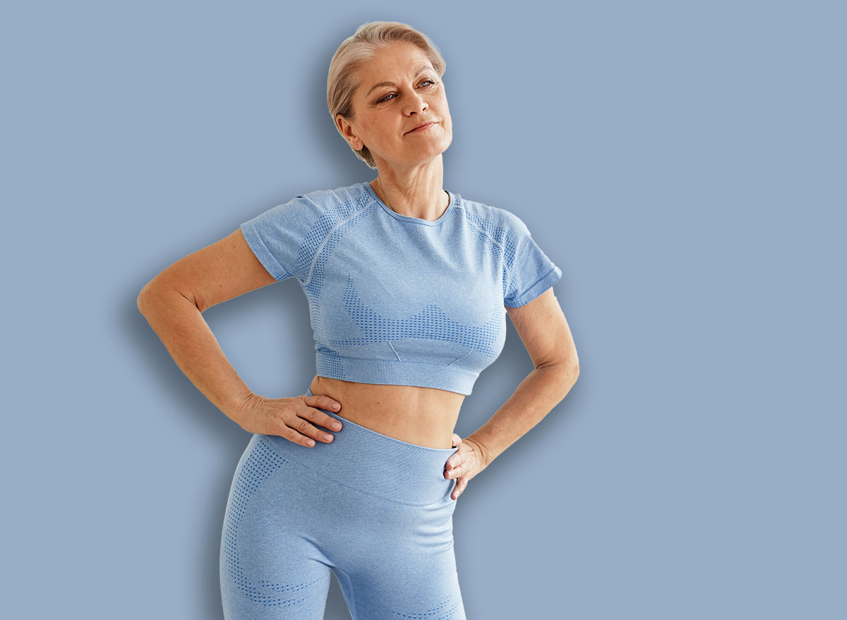 Aged Caucasian woman standing with hands on hips stretching in living room, wearing athletic outfit, looking away from camera, appearing focused and confident