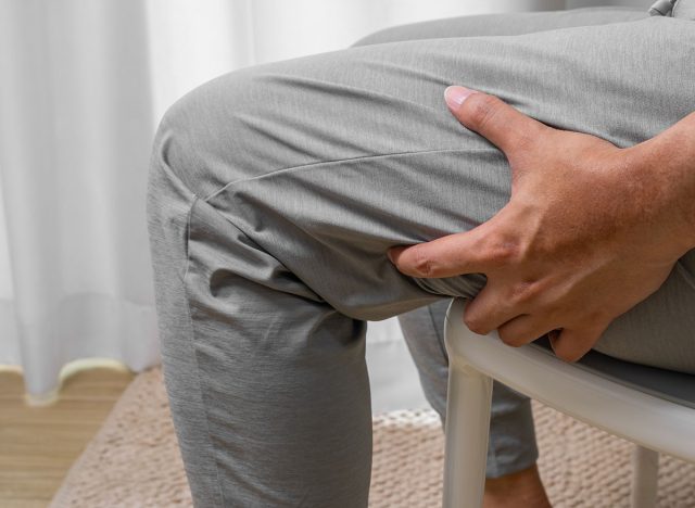 Man, indoors gripping painful hamstring post-exercise, suggests muscle strain, fatigue, or soft tissue injury from overuse or poor warm-up.