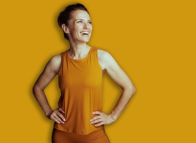 An excited woman in vibrant yellow activewear and leggings poses with hands on hips, embodying an optimistic and active lifestyle. The warm background enhances her joyful mood.