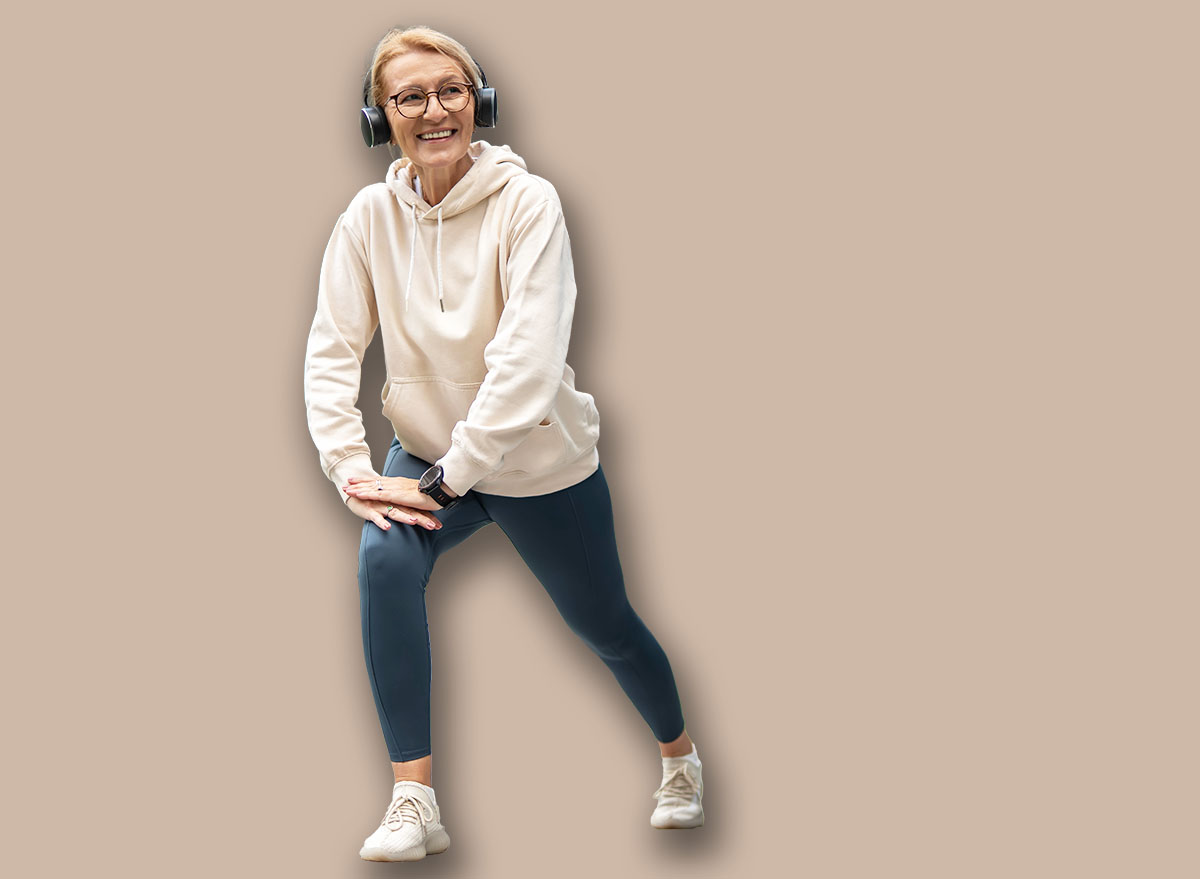 Smiling senior woman in headphones stretching her legs on a paved walkway, exercising outdoors in an urban environment with modern buildings in the background, promoting active aging and well-being