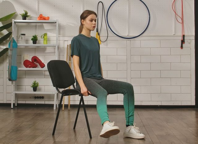 A young woman performs a resistance band exercise while seated on a chair in a fitness studio.