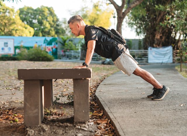 Middle aged Southeast Asian man doing incline pushups on a concrete bench while wearing a loaded rucksack for resistance training.