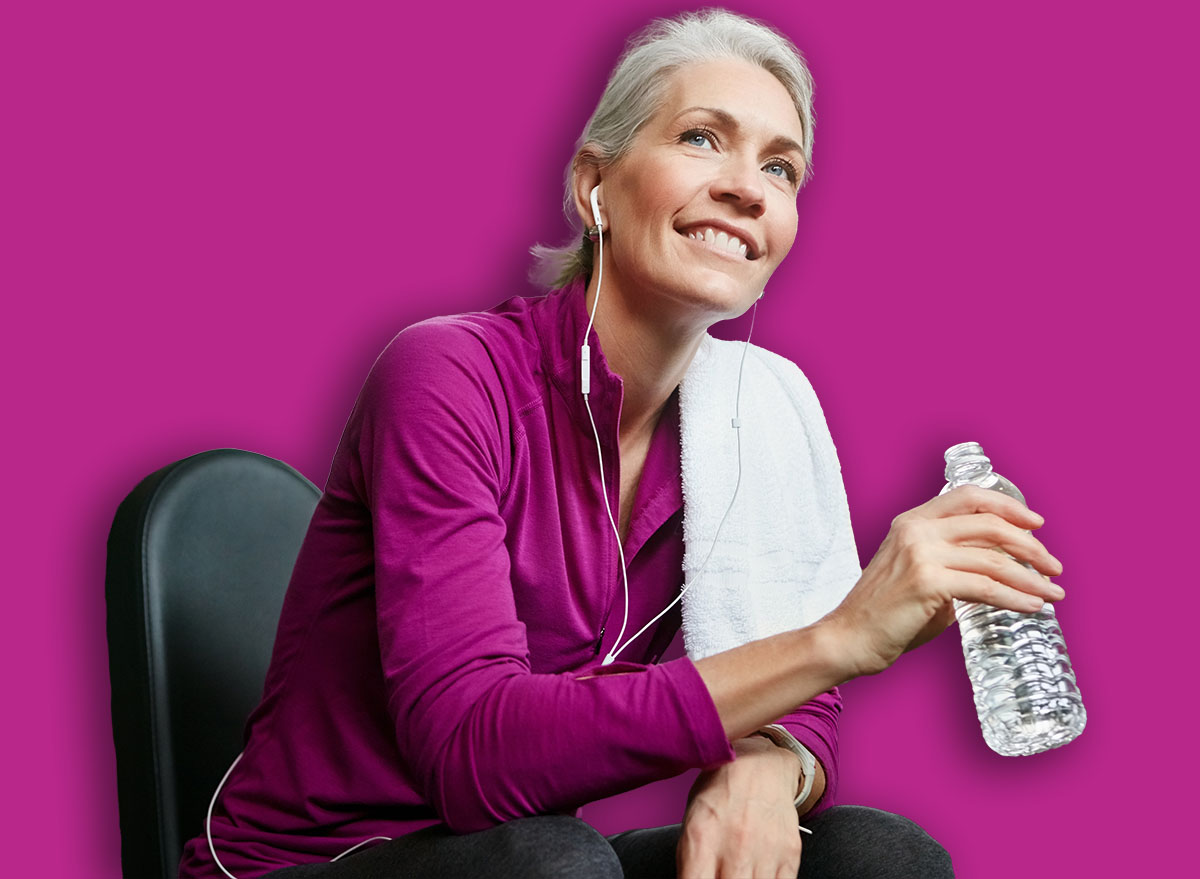 Mature, happy woman and relax with bottle water, earphones or music on machine for workout or exercise at gym. Female person with smile for break, mineral beverage or thirst in rest at health club