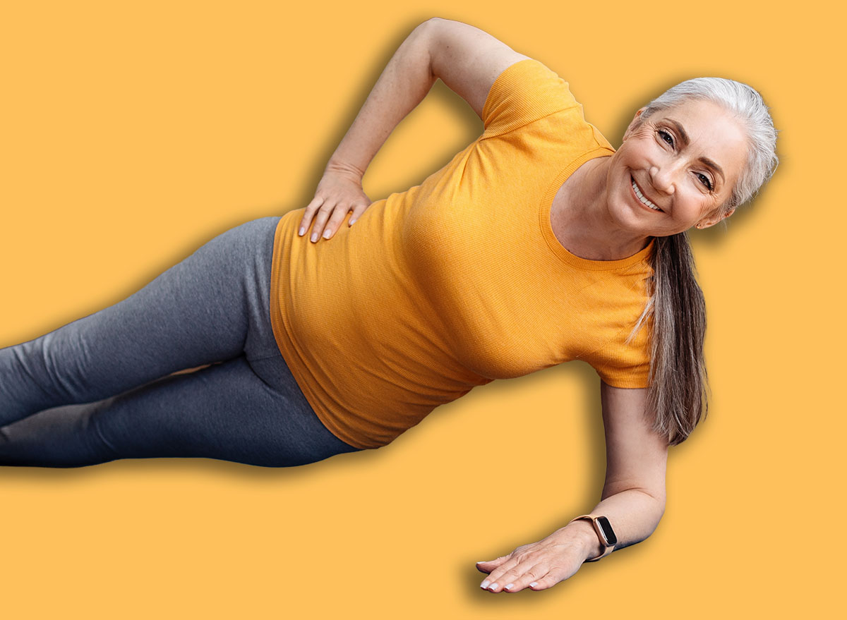 Happy fit senior woman standing in side plank, training on yoga mat in living room, using laptop, watching tutorial or working out with trainer