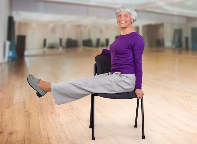 Happy charming beautiful elderly woman is doing exercise on a chair. Exercising gymnastics for health in the fitness room. Sports training.
