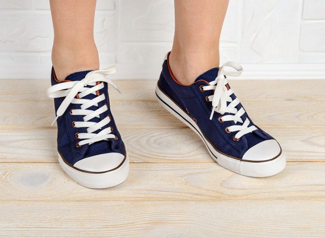 Woman feet shod in classic bllue gumshoes with white shoelaces and standing on toes on a white wood floor. Stand on tiptoe in sports shoes close-up. Front view.