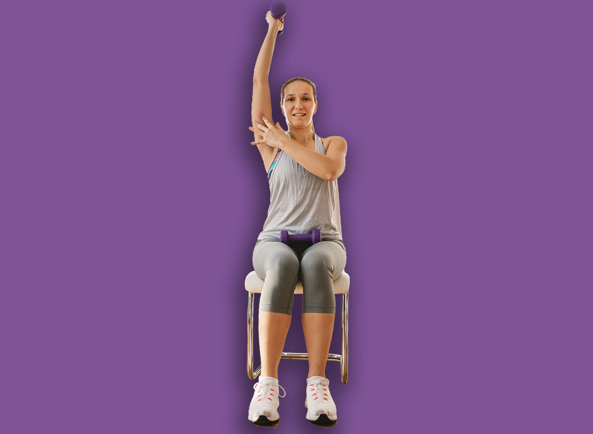Muscular sportswoman in shape sitting on the chair at home, lifting dumbbell and showing biceps with other hand.