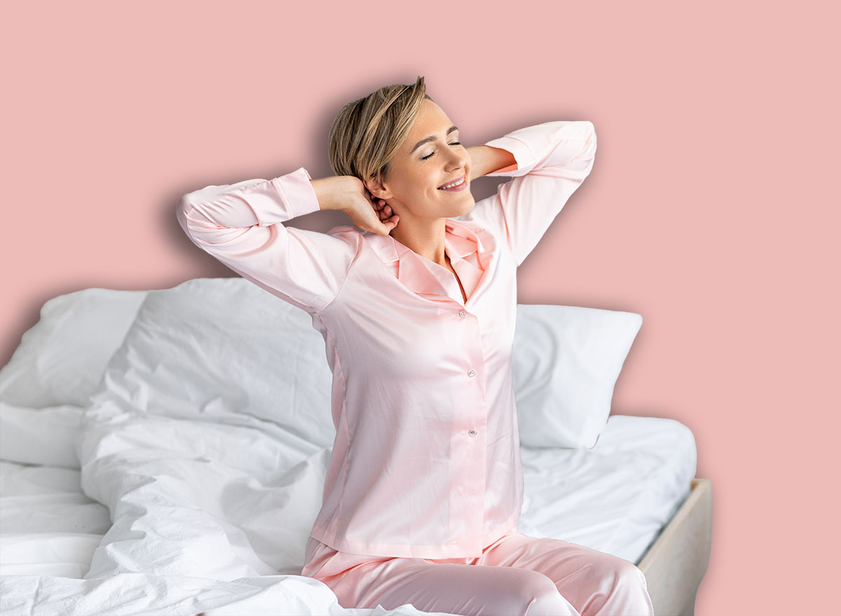 Good Morning. Cheerful positive middle-aged lady stretching arms and back after waking up, happy female in pink silk sleepwear sitting on bed, feeling great, enjoying good start of the day, free space