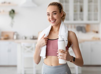 Covid-19 lockdown, body and health care, thirst and coach recommendations. Cheerful young pretty blonde lady in sportswear with towel on shoulders holds bottle of water in minimalist kitchen interior