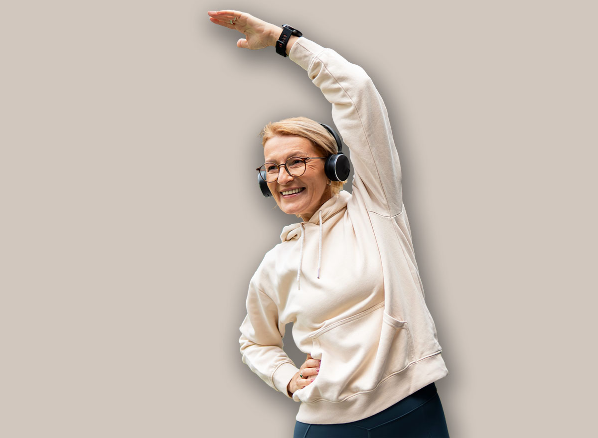 Senior woman in athletic wear and headphones doing side bends in a modern urban park, smiling confidently while stretching during a morning fitness routine and enjoying outdoor wellbeing