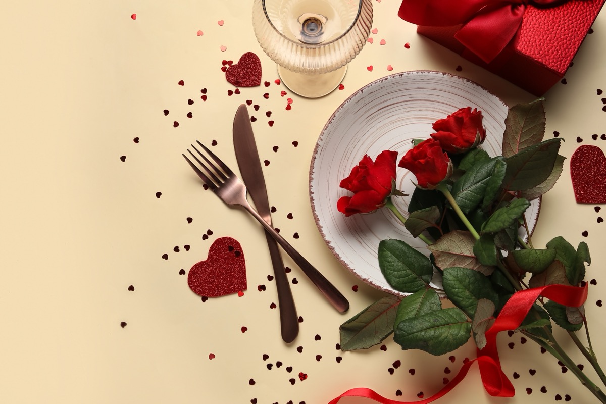 Beautiful table setting with gift box, hearts and red roses on yellow background. Valentine's Day celebration