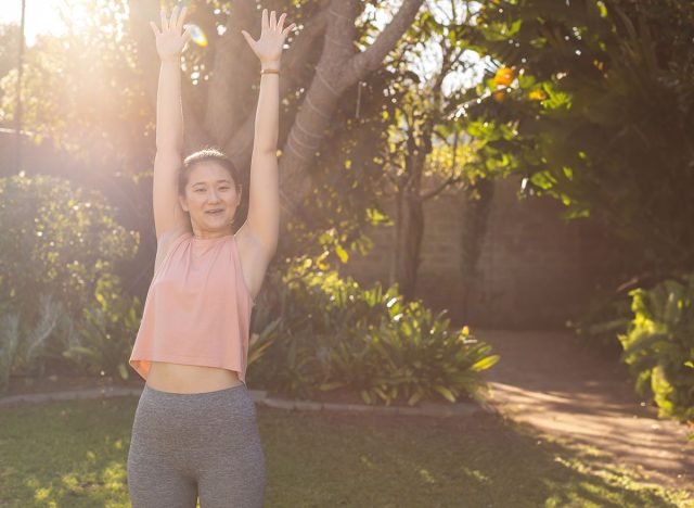 Asian woman stretching arms overhead on grassy lawn in residential backyard, wearing smartwatch. Fitness, activewear, wellness, flexibility, mindfulness, home workout, outdoor