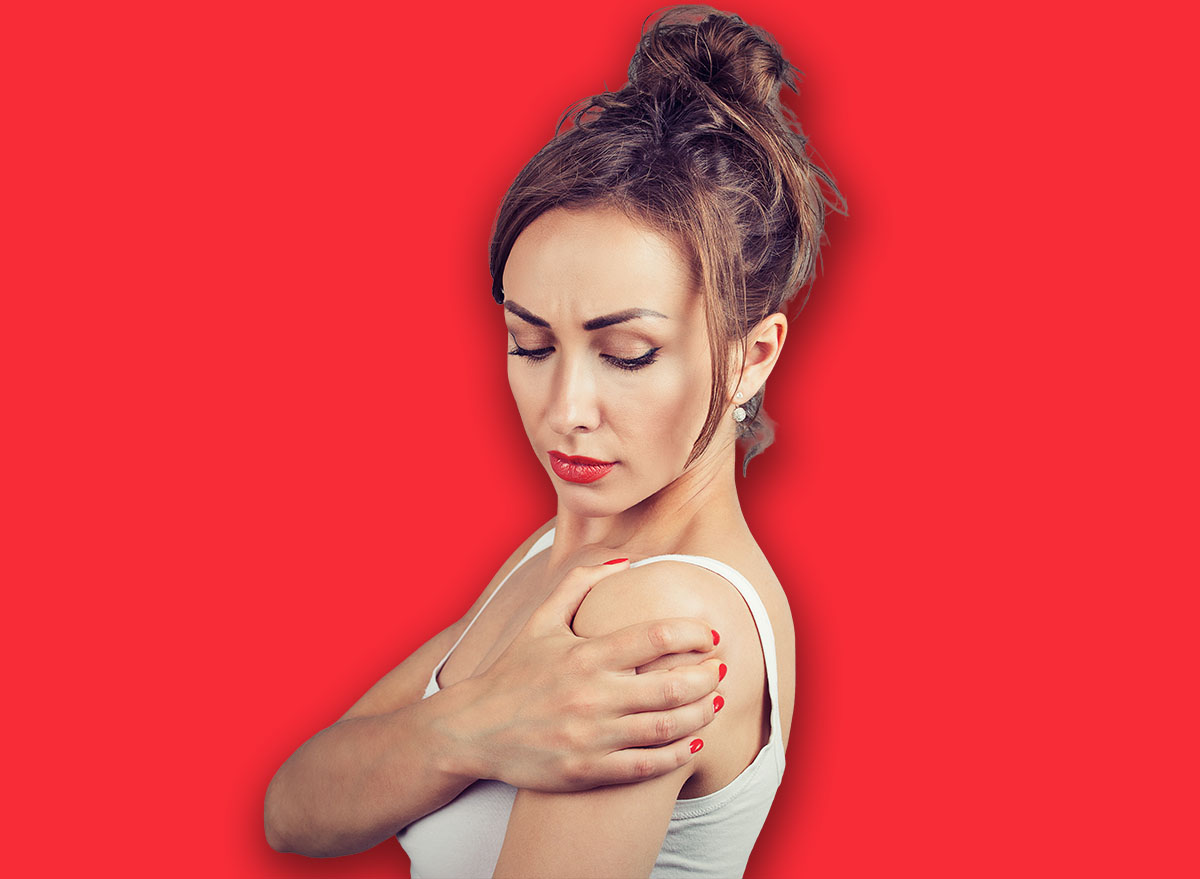 Young brunette rubbing shoulder having pain in joint and standing on gray background