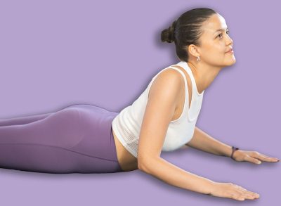 Focused young Asian woman performing stretching and back bending asana Salamba Bhujangasana, or Sphinx Pose, during group yoga training in sunlit fitness studio