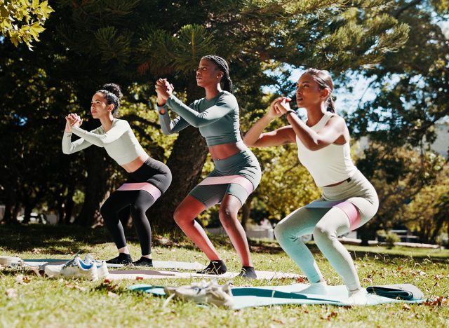 Women, squat or yoga class with pilates in nature for exercise or balance at outdoor park. Active group, female people or yogi friends with resistance band on mat for fitness, health or wellness