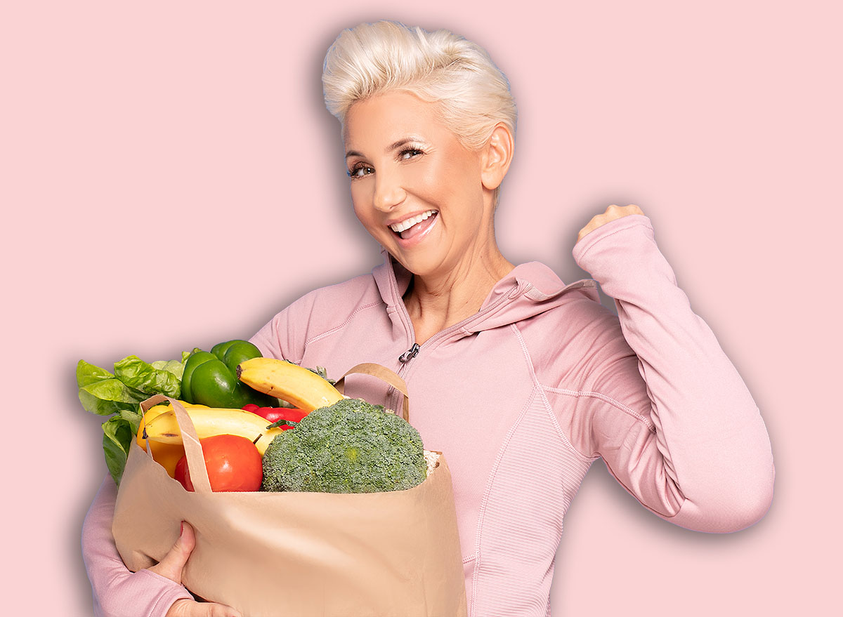 Smiling adult woman holding shopping bag full of groceries posing on blue studio background. Healthy food shopping. Paper package with vegetables and fruits, happy female buyer came from market.