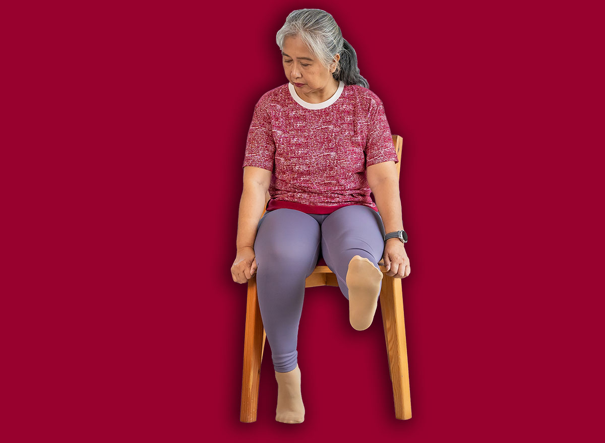 Senior woman sitting on a wooden chair, raising leg to stretch muscles and knees, Training exercise online with tablet In Living Room During Quarantine