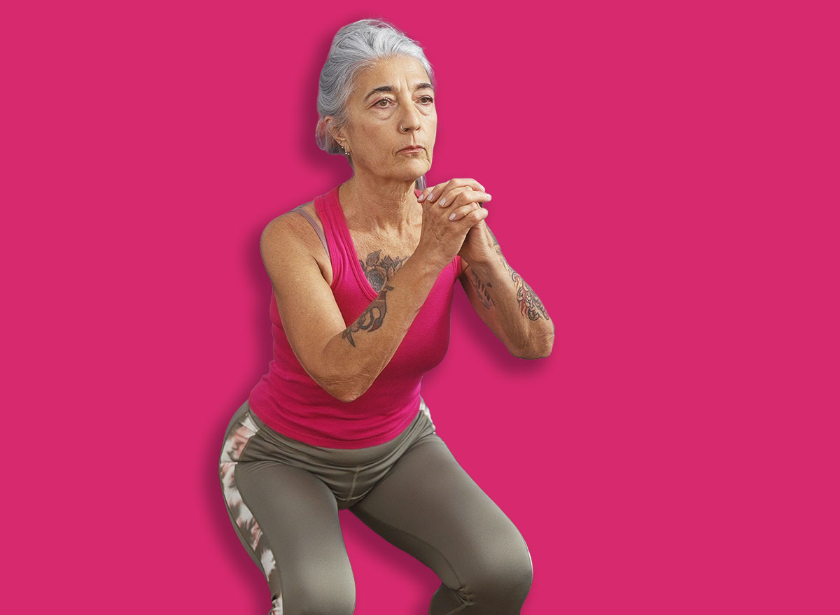 Grey-haired woman exercising at gym performing squats in an indoor sport center with gym equipment visible in the background