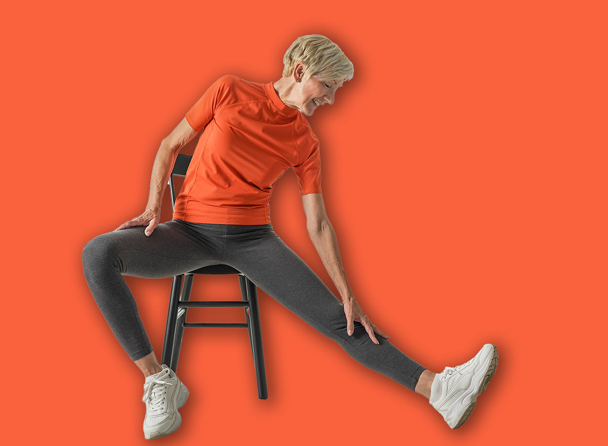 An elderly woman in an orange shirt and gray leggings sits on a chair, stretching her leg to the side. She reaches toward her foot in a bright living room with plants, promoting senior wellness