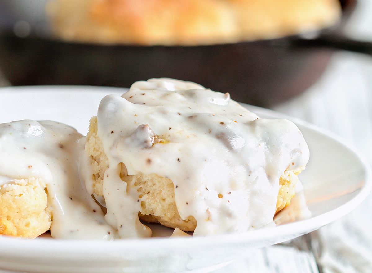 American biscuits from scratch covered with thick white sausage gravy. Selective focus with cast iron skillet / pan in the background over a white table.