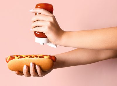 Woman adding ketchup onto tasty hot dog on pink background