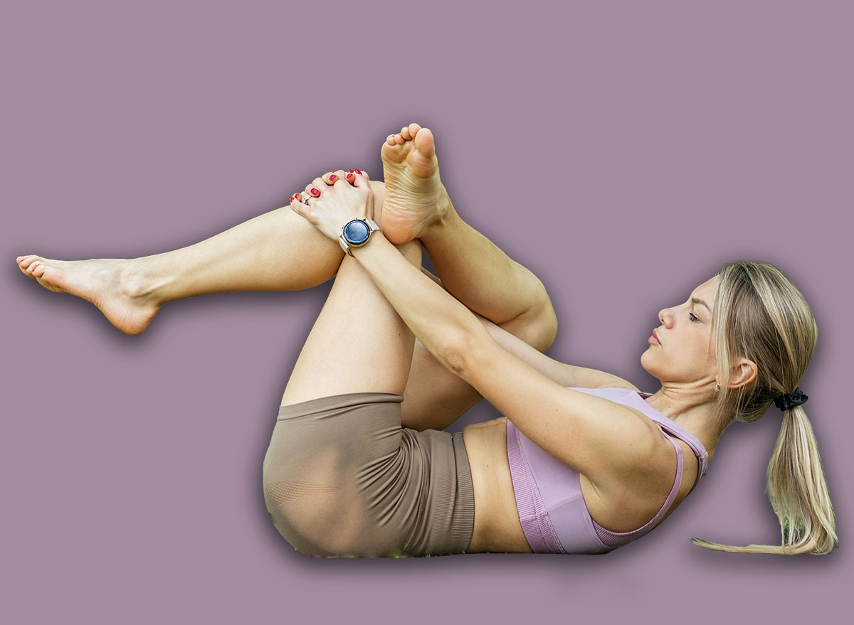 Young fit woman performing a figure four stretch on her back in a peaceful forest park, targeting hips and glutes flexibility.