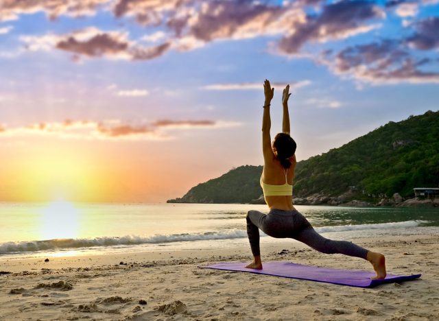 A woman practices yoga on a sandy beach at sunrise, poised in a high lunge with her arms reaching toward the sky.