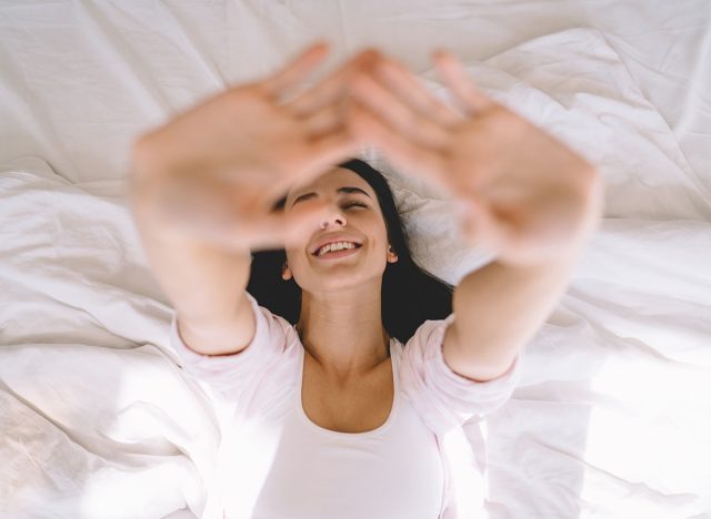 Top view of happy female with eyes closed lying on back on cozy comfortable bed and stretching hands up while relaxing free time at home
