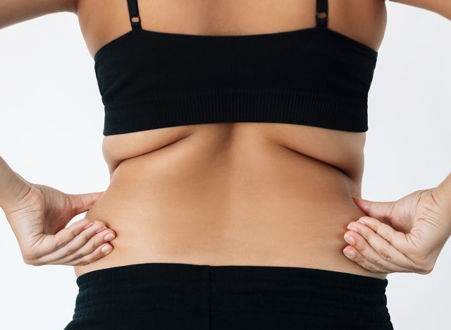 Cropped shot of young woman standing with her back holds fat folds on her sides isolated on a white background. Overweight, flabby and sagging muscles, rolls on lower back. Excess weight