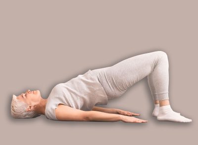 A content senior woman with short hair lies on her back carrying out a bridge exercise on a yoga mat in spacious, well-lit living room. She wears comfortable attire and has pair of dumbbells nearby