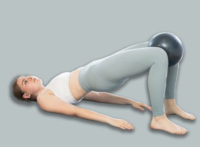 Young woman performs glute bridge with softball in knees during Pilates training. Ball is clamped in knees and pelvis is raised and lowered