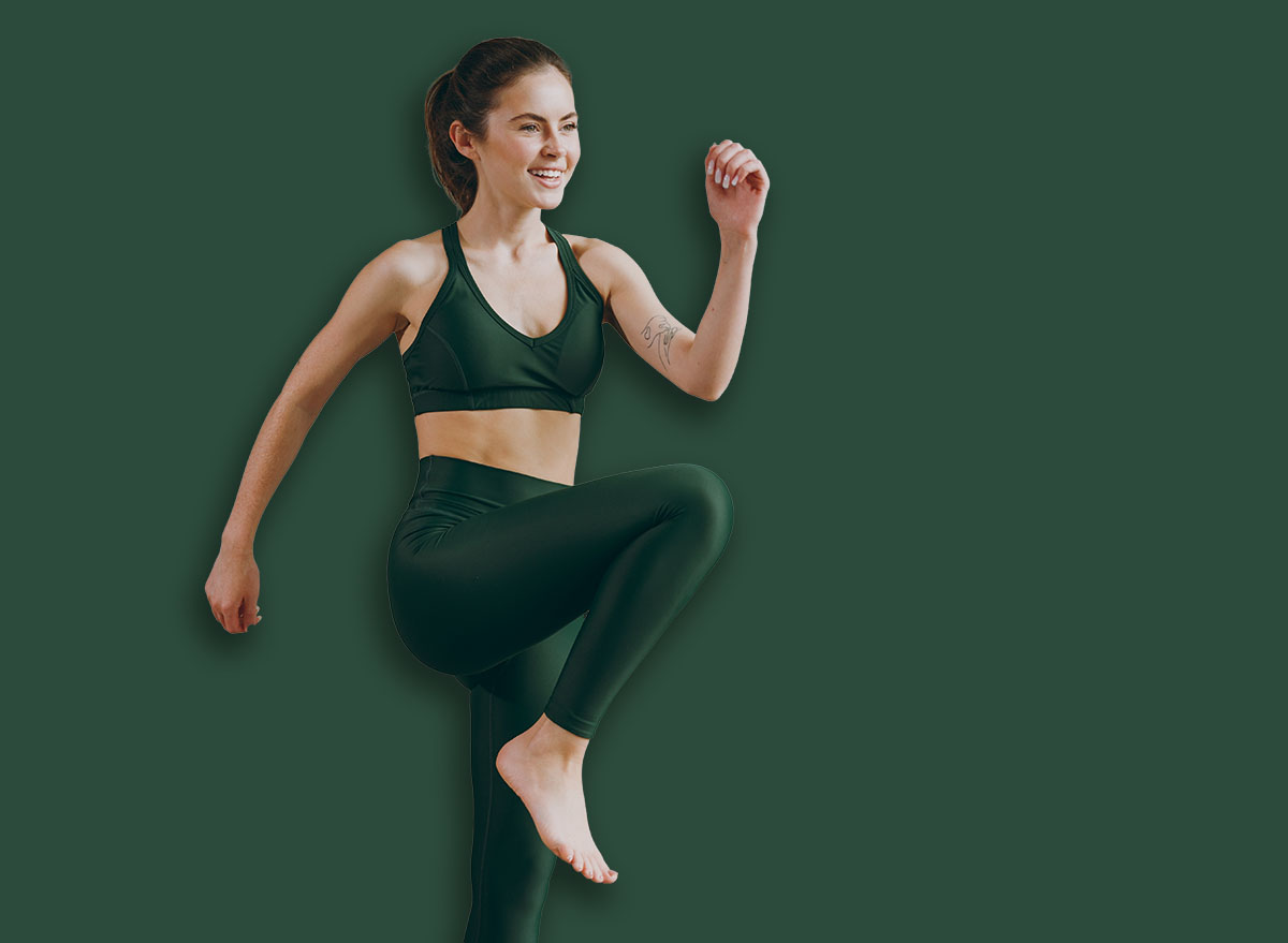 Side profile view young strong sporty athletic fitness trainer instructor woman wear green tracksuit raise up leg run in place training do exercises at home gym indoor Workout sport motivation concept