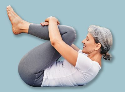 Side view of mature woman in sports wear lying on mat hugging her knees to chest after yoga practice outdoor in park, healthy active senior female doing stretching exercises in morning on fresh air
