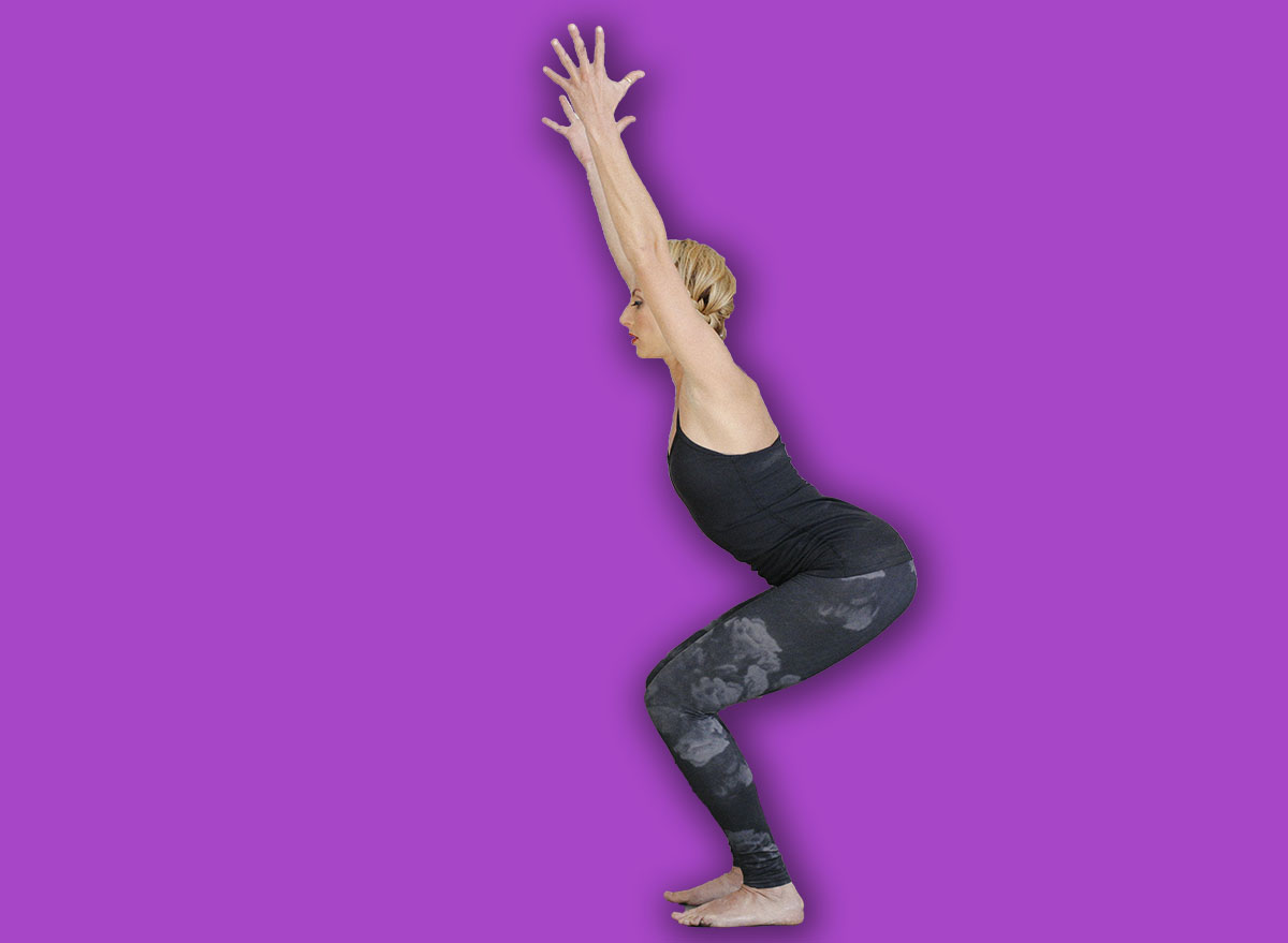 A blonde woman in a black leotard and leggings, standing on a yoga mat in a room, doing yoga, squatting down with her arms raised