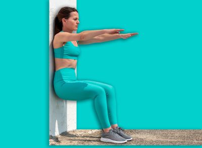 Sporty woman doing squats against a white wall outside in the sun.