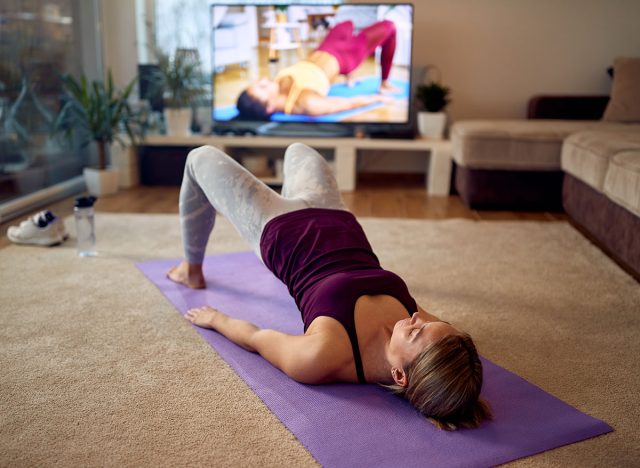 Female athlete doing glute bridge exercise on the floor in front of a TV at home.