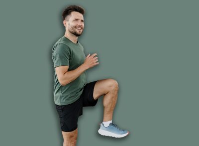 A man warms up under a bridge, lifting his knee high in a dynamic stretching exercise.