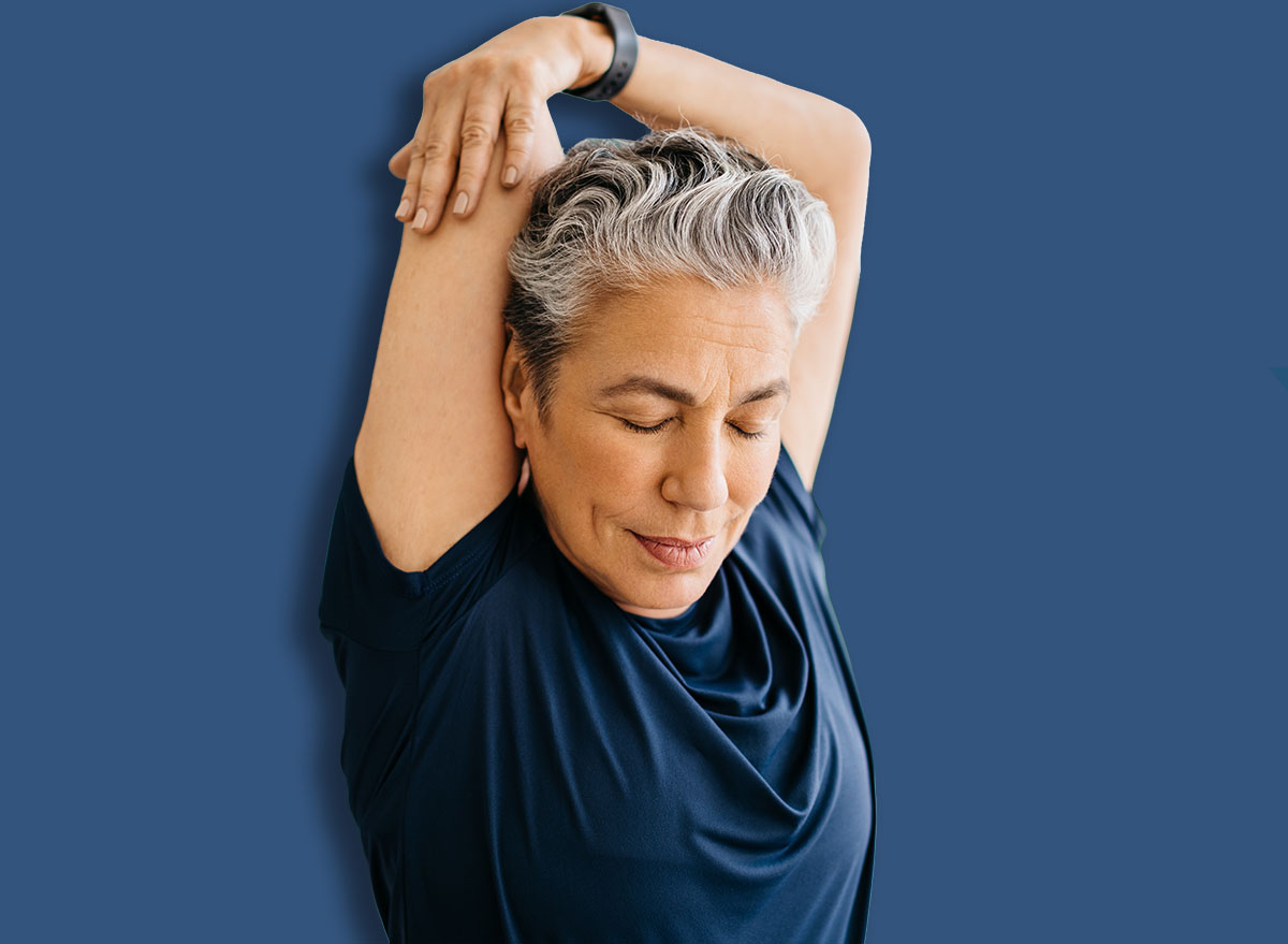 Senior woman doing an overhead arm stretch for a healthy tricep in a peaceful home workout session. Active woman with grey hair practicing yoga for a better physical wellbeing in her retirement age.