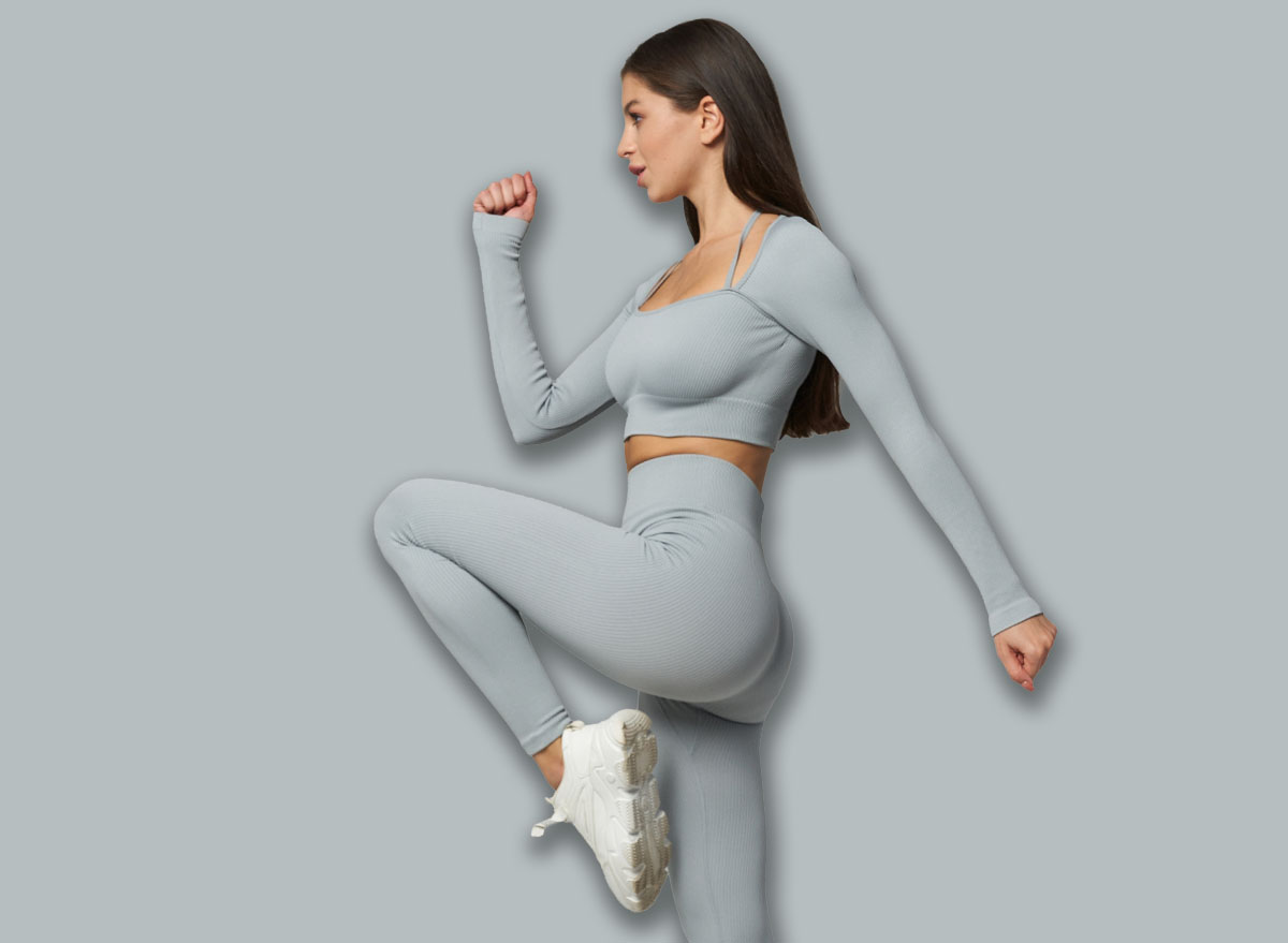 Young woman in gray workout attire performing a high-knee exercise, raising her right knee with arms bent, showcasing fitness, balance, and active lifestyle against a plain setting