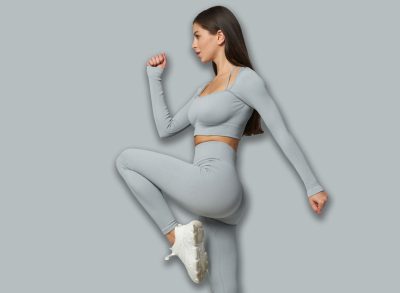Young woman in gray workout attire performing a high-knee exercise, raising her right knee with arms bent, showcasing fitness, balance, and active lifestyle against a plain setting