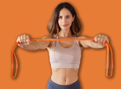 A woman is performing a home workout using a resistance band. She shows determination while working out in a well-lit living space with wooden flooring and shelves in the background.
