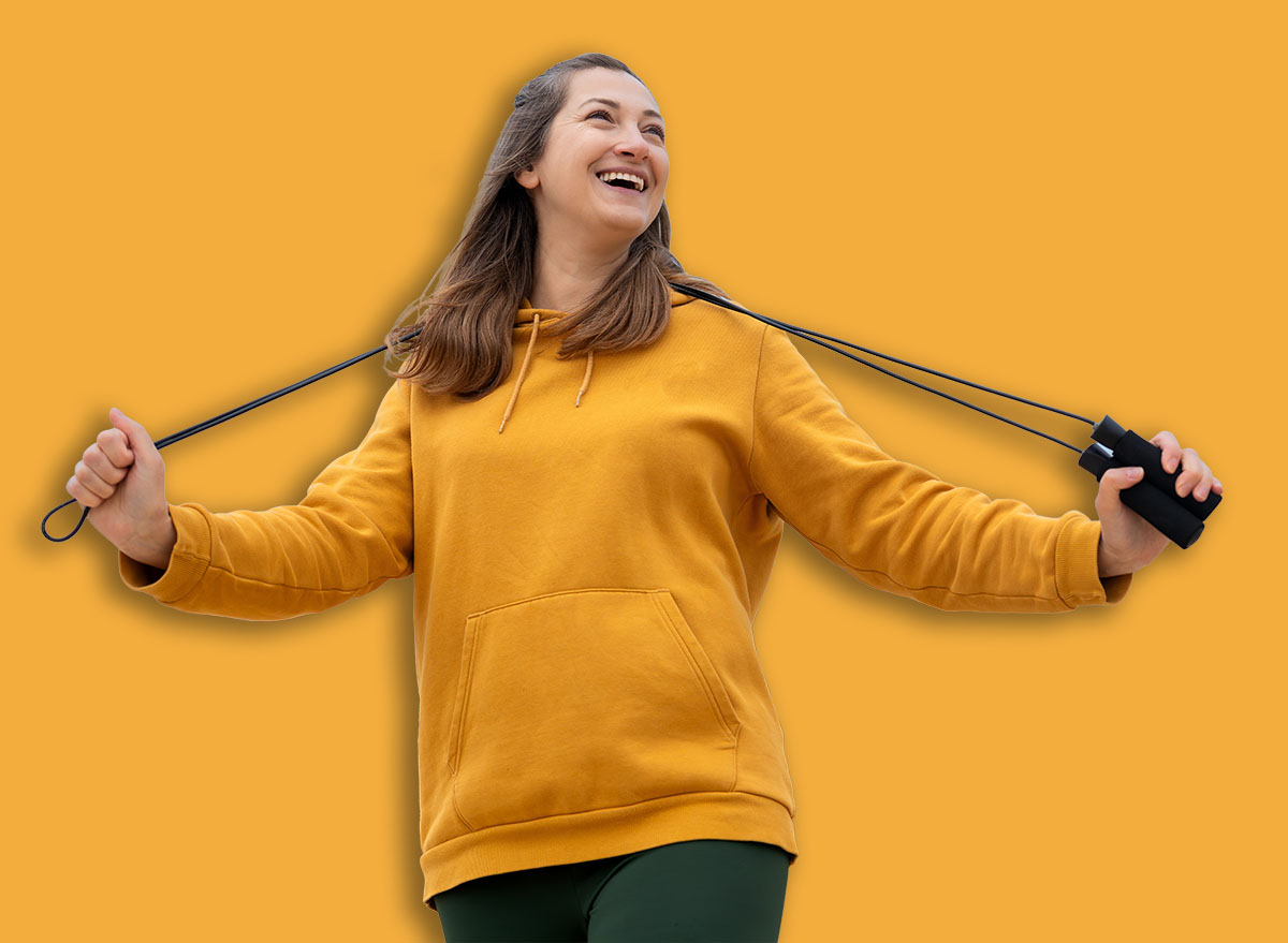 A woman in a yellow hoodie holds a jump rope and smiles against a sky background on the stadium