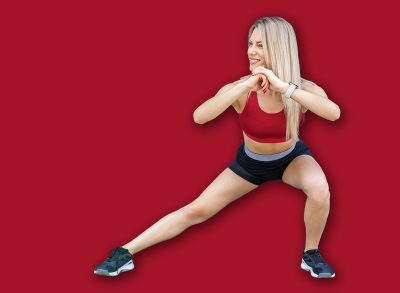 Fit woman in red sports bra and black shorts doing side lunges outdoors against modern brick wall background.