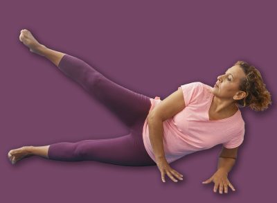 A mature hispanic woman exercises indoors on a blue mat in a gymnasium, demonstrating flexibility and strength as she performs a leg lift in a well-equipped sports center.
