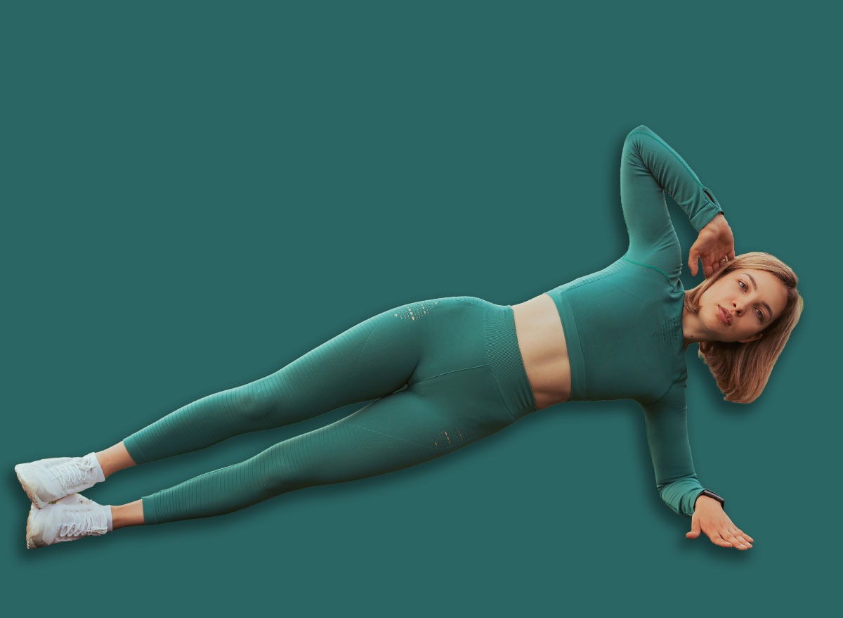 Fit sportswoman in blue activewear doing side plank exercise while balancing on green field at stadium during workout in summer and looking at camera