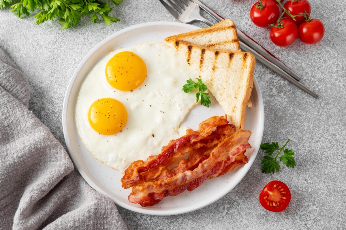 Breakfast with fried eggs, bacon and toasts on a white plate with cup of coffee on a stone background. Top view, selective focus
