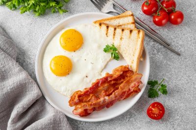 Breakfast with fried eggs, bacon and toasts on a white plate with cup of coffee on a stone background. Top view, selective focus