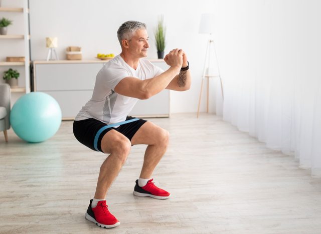 Morning training. Sporty mature man doing squats, exercising in living room interior, copy space. Athletic sportsman having domestic training during covid-19 lockdown