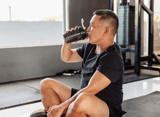 Southeast Asian man in his 50s sits on gym mat, drinking water or sports drink post ab workout, recovering and hydrating.